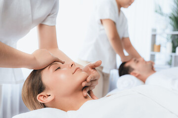 Caucasian couple enjoying relaxing anti-stress head massage and pampering facial beauty skin recreation leisure in dayspa modern light ambient at luxury resort or hotel spa salon. Quiescent