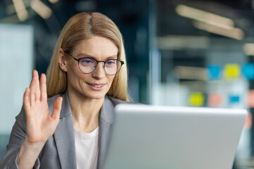 Professional woman in office using laptop for video call. She waves hand at screen, engaged in virtual communication. Glasses and suit reflect work environment. Technology enhances teleconference.