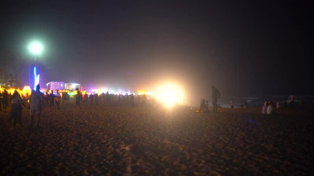 Slow panning shot showing crowd of people enjoying the night at a beach in goa maharastra with brightly lit shacks in the distance showing the vibrant nightlife