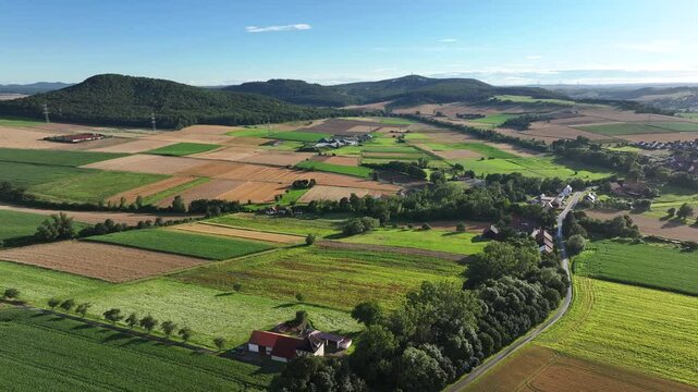 Aerial view of patterned fields and a quaint village surrounded by hills and trees in summer, Humme, Hessia, Germany.