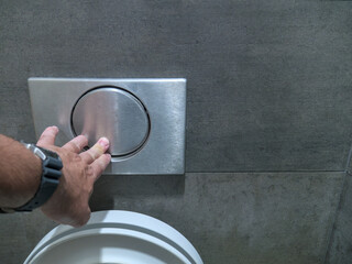 Close-up shot of a Caucasian man's hand pressing a large button on the wall to flush the toilet bowl in a public restroom