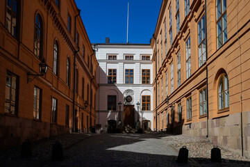 A peaceful cobblestone alleyway surrounded by warm-toned historic buildings captures the midday sunlight. The clear blue sky enhances the serene atmosphere, inviting exploration.