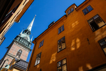 A historic Riddarholmen church with a tall spire is framed by buildings in a cobblestone alleyway at Gamla stan in Stockholm. The bright blue sky enhances the picturesque atmosphere of the scene.