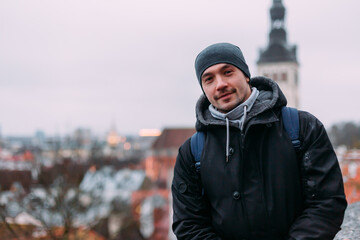 man standing on tallinn viewpoint with cityscape background, winter clothing and beanie hat, smiling traveler in old town, scenic urban landscape, relaxed outdoor portrait