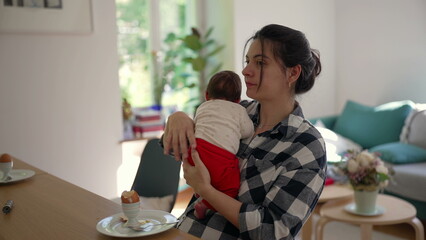 Mother holding newborn baby while seated at a table, demonstrating the love and challenges of...