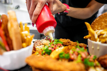 A close-up of a hand squeezing ketchup onto a plate of fast food, including fries and fried chicken, garnished with fresh herbs. Perfect for illustrating casual dining or fast food themes.
