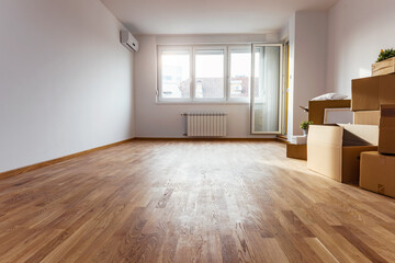An empty apartment with wooden flooring, large bright windows, and several stacked brown cardboard boxes in the corner, suggesting a recent move or home renovation. The space feels airy and new.