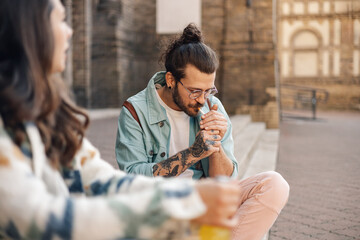 Young tattooed hipster guy sitting on street and lighting a cigarette.