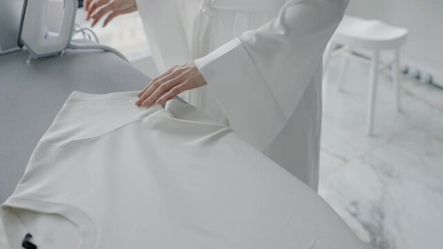 A woman irons a white shirt on an ironing board in a clean, minimalist setting, showcasing domestic chores. Woman Ironing Clothes at Home in Bright Modern Apartment