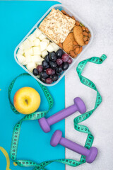 Healthy snack, diet, fitness, weight control. Lunch box with a light snack, cereal cookies, fruits, berries, nuts, dumbbells and a tape measure on a fitness mat.