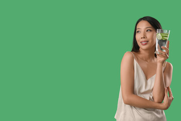 Young Asian woman with glass of cucumber water on green background