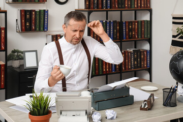 Displeased mature man with cup of coffee and vintage typewriter sitting at table in office