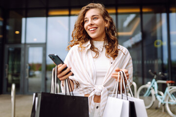 Young woman enjoying shopping while checking her phone outside a store on a sunny day in a vibrant urban area. Consumerism, sale, black friday. Online shopping.