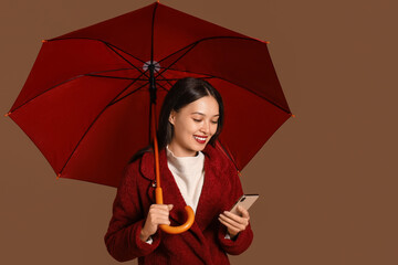 Beautiful young Asian woman with umbrella and mobile phone on brown background