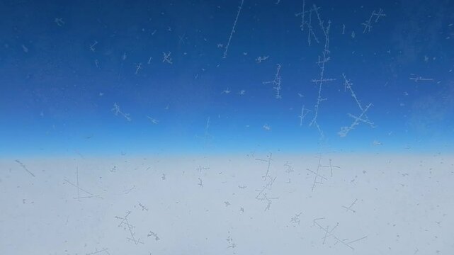 Flight above white clouds and horizon of clear blue sky. Aerial view through window with white traces of frost due to very low temperature at high altitude during flight. Topics: weather, meteorology