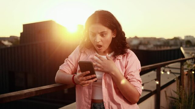 Amazed young indian woman got notification message and looking at screen with puzzled face at home terrace Shocked beautiful female reacting to news and feel confused with sunset on background