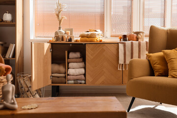 Open commode with stacks of stylish clothes and autumn  decorations near window in living room