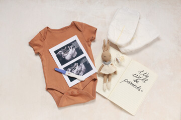 Baby clothes, sonogram images, pregnancy test and notebook with text WE WILL BE PARENTS on grunge beige background