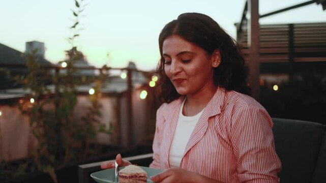 Pretty young indian woman looking around to make sure nobody watching and eating cake with great pleasure at home terrace Smiling female biting and enjoying bakery indoors Break the diet concept