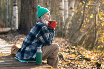 Satisfied middle aged woman spending tranquility time alone walking in autumn forest dreaming drinking hot thermos tea. Relaxed pleased female rest on eco path enjoys calm late fall park, stress free.