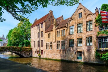 Naklejka premium A stunning view of a canal in Bruges, Belgium. The calm water reflects the vibrant buildings along the banks. Lush green trees frame the scene under a blue sky with soft clouds.