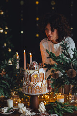 Beautiful woman in knitted sweater making handmade homemade delicious gingerbread house for family Christmas celebration. Elegant table setting. Cozy home atmosphere, festive mood, candlelight