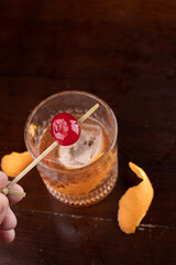 close up of old fashioned cocktail cherry with bourbon translucent ice in fancy glass orange peel on wooden table