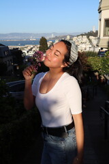 Chinese girl holding flower and posing at top of Lombard Street area in San Francisco with the city in the back during golden hour sunset