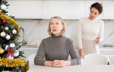 Sad old woman sitting at table while middle-aged female family member scolding her in the kitchen...