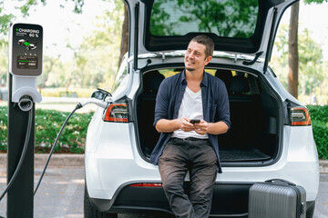 Man using smartphone online banking application to pay for electric car battery charging from EV charging station during vacation holiday road trip at national park or summer forest. Exalt