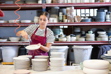 Young woman worker in uniform paints blanks of ceramic products at factory