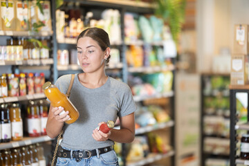 Interested young girl in casual gray t-shirt and jeans holding and examining organic juice bottles,...