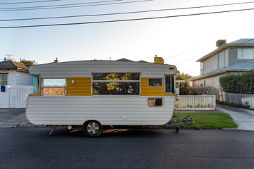 An old vintage caravan with a retro design parked on a suburban street in front of a typical Australian suburban house with a picket fence.