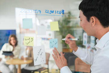 Business man uses a glass wall covered with sticky notes to outline processes during meeting. Engaged colleagues gather around a glass brainstorming board. plotting ideas and strategies.