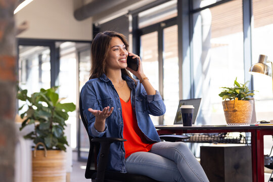 Talking on smartphone, woman sitting at desk with laptop and coffee in office