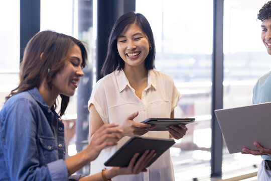 Smiling diverse colleagues discussing project, holding tablets and laptop in modern office