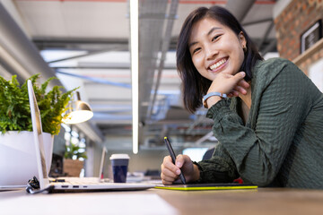 Smiling asian woman using digital pen and tablet while working in modern office
