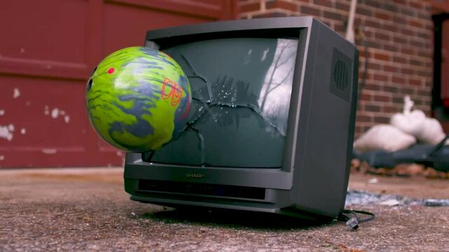 Old vintage black tv with being thrown with a colorful blue and green bowling ball in slow motion during day time with a brick wall and metal door in the background. Screen gets cracks and is broken.