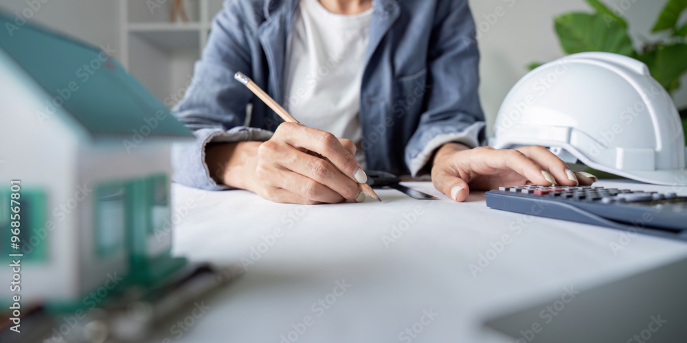 Poster engineer working on architectural plans with calculator and model house