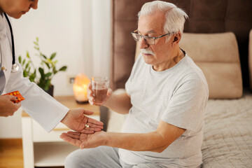 Senior man sitting on bed in bedroom and his doctor giving him painkillers.