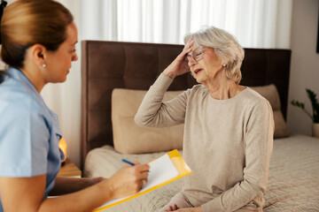 Ill senior woman with migraine sitting on bed and telling symptoms to nurse during her checkup.