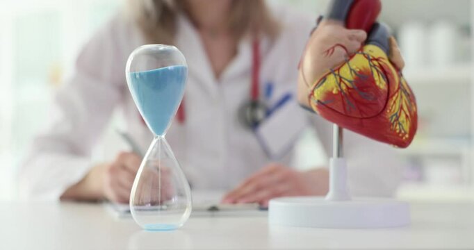 Cardiologist studying anatomical model of heart and observing hourglass symbolizing importance of heart treatment time