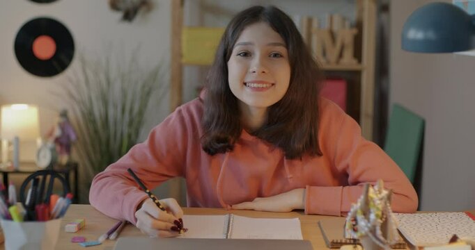 Portrait of cheerful teenage girl doing homework writing then looking at camera sitting at desk in bedroom. Education and teenager concept.