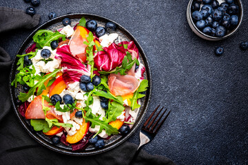 Foodie food salad with peach, jamon, soft cheese, blueberries and arugula on black background, top view