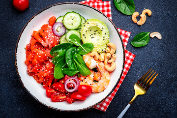 Seafood salad bowl with salmon, shrimp, avocado, spinach, cucumber, tomato, cashew and sesame. Black background, top view
