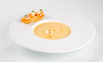 A bowl of creamy soup with croutons served in a white plate, accompanied by a slice of bread topped with cheese and herbs on the side.