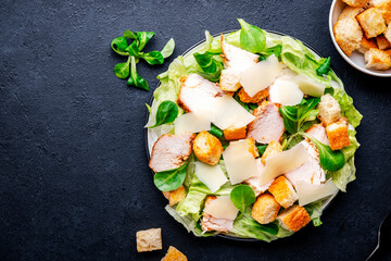 Caesar salad with grilled chicken slices, iceberg lettuce, croutons, parmesan cheese and classic dressing. Black background, top view