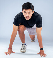 An Asian man in a ready-to-sprint stance, wearing a black t-shirt, gray shorts, and athletic shoes,...