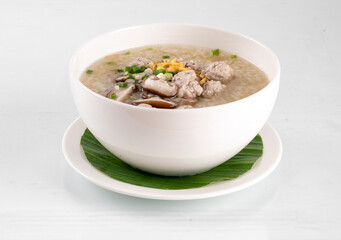 A delicious bowl of congee with garnish, served in a white bowl and placed on a white saucer with a green leaf mat on a light background.