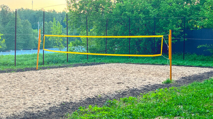 A yellow volleyball net is set up on a sandy field. The net is attached to a wooden pole. The field is surrounded by trees and grass